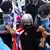 A woman with while hair partially wrapped in the UK flag is gripped under the arms by two police officers who are taking her away