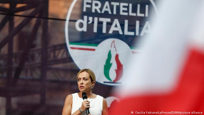 Giorgia Meloni, party logo in the background that reads Fratelli d'Italia with stylized flames 