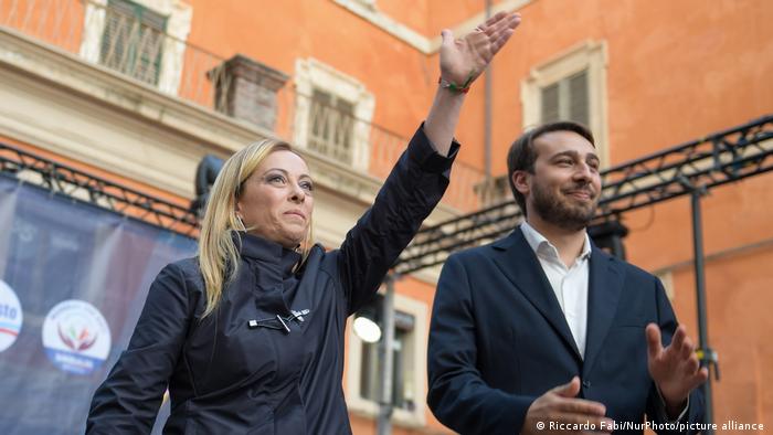 Giorgia Meloni outdoors, standing next to a man who is applauding, she is holding up left arm. 