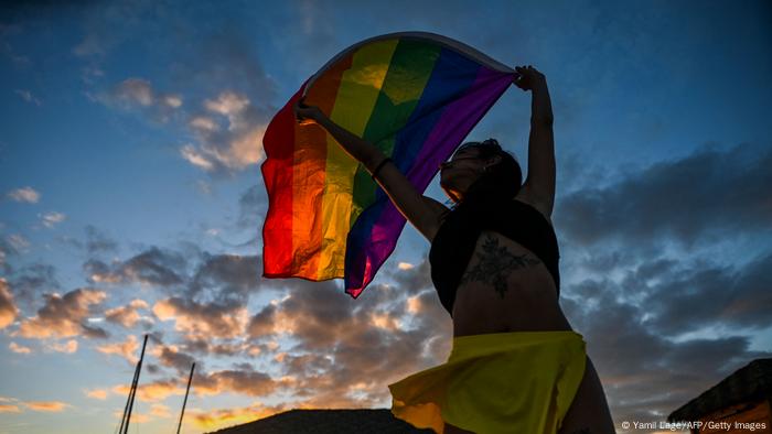 A dancer, photographed from below, holds up a rainbow flag against an evening sky A dancer, photographed from below, holds up a rainbow flag against an evening sky