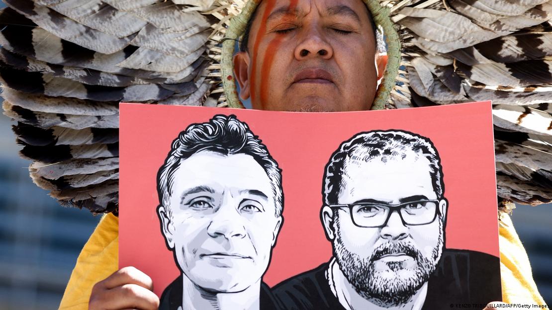 Foto de indígena com cocar segurando imagem com desenho dos rostos de Dom Phillips e Bruno Araújo