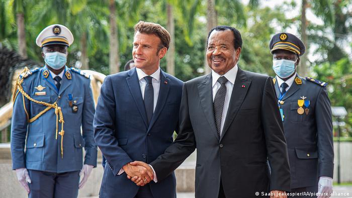 French President Emmanuel Macron (l) and Cameroonian President Paul Biya (r) shake hands in the capital Yaounde as two uniformed men stand behind them 