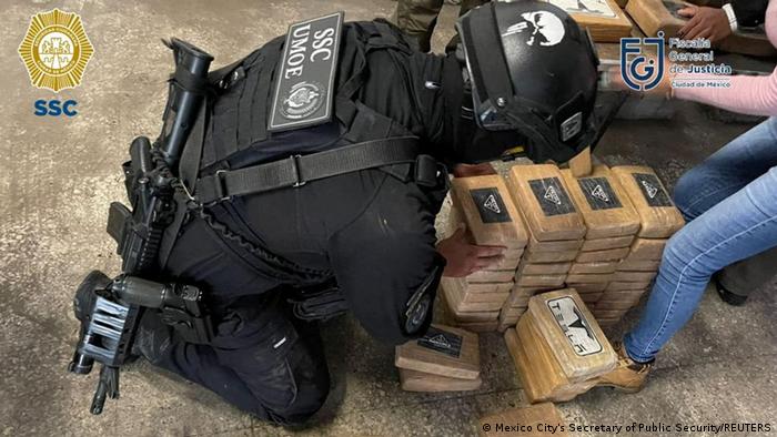 A police officer in heavy-duty black uniform and black helmet, a rifle slung across his back, bends over a stack of brick-like, pale brown packages wrapped in plastic.