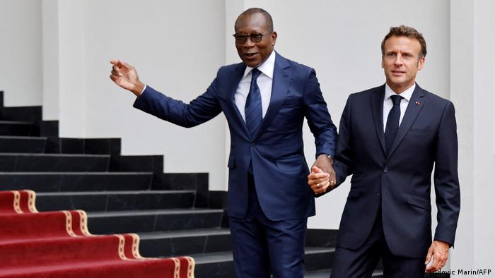 Patrice Talon's holds Emmanuel Macron's wrist and gestures as the two stand on stairs 