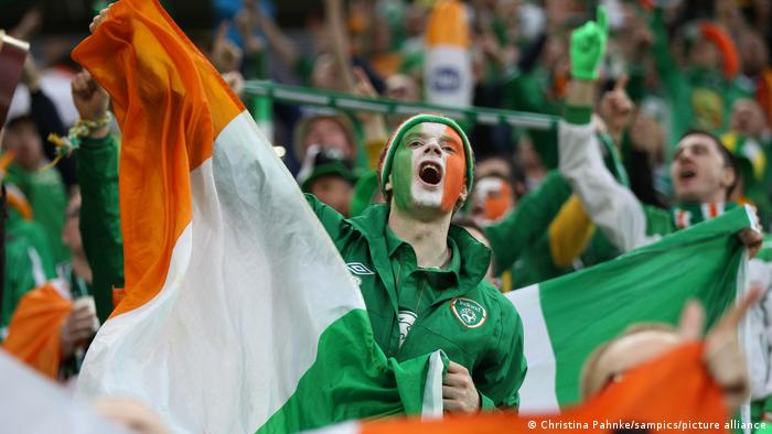 Ein Fußballfan mit grün-weiß-orange geschminktem Gesicht schwenkt eine irische Flagge und singt, im Hintergrund die Zuschauerränge mit singenden Fans.
