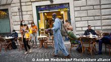 A street vendor tries to sell his goods in front of a cafe', in Rome, Monday, Oct. 26, 2020. For at least the next month, people outdoors except for small children must now wear masks in all of Italy, gyms, cinemas, swimming pools, and movie theaters will be closed, ski slopes are off-limits to all but competitive skiers and cafes and restaurants must shut down in early evenings, under a decree signed on Sunday by Italian Premier Giuseppe Conte, who ruled against another severe lockdown despite a current surge in COVID-19 infections. (AP Photo/Andrew Medichini)