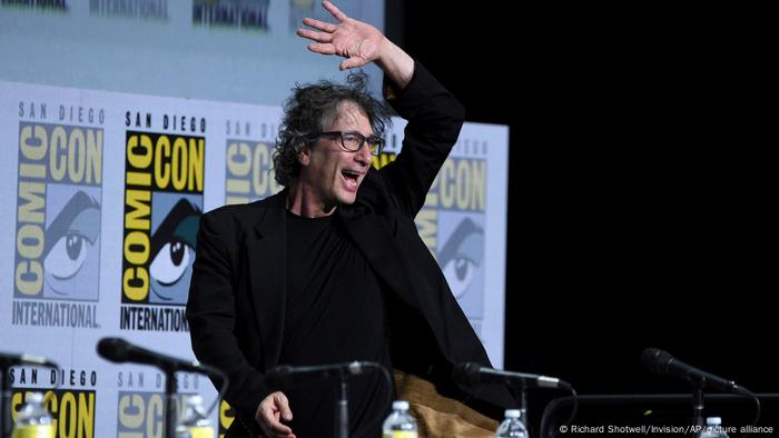 Neil Gaiman hebt die Hand und winkt einem Publikum auf der ComicCon in San Diego zu. Neil Gaiman hebt die Hand und winkt einem Publikum auf der ComicCon in San Diego zu.