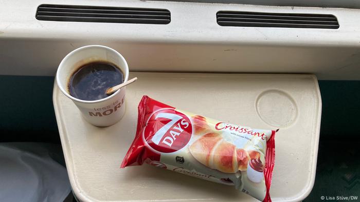 A cup of coffee in a paper cup and a croissant in a bag on a train table.