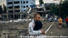 Ein Paar steht neben zerstörten Gebäuden und umarmt sich. Lokalen Medien zufolge wurden durch russischen Beschuss ein Supermarkt, ein Hochhaus und eine Apotheke beschädigt. +++ dpa-Bildfunk +++