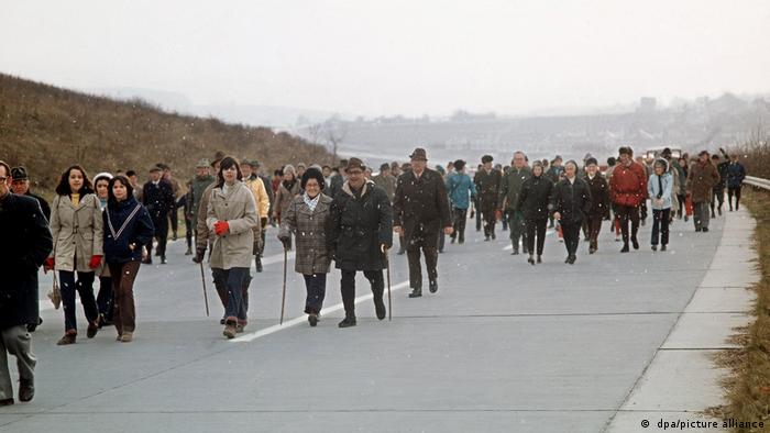 A large crowd of people in winter clothes, some with walking sticks, walking along a three-lane highway.