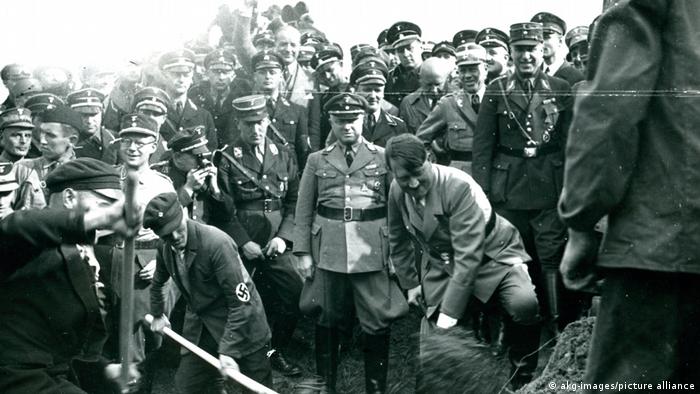 Black and white photo of a crowd of men in Nazi uniforms standing behind Hitler and another man with a swastika armband, both of whom are digging at the earth with spades.