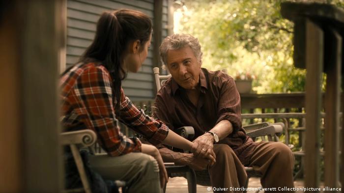 Dustin Hoffman sitzt in As They Made Us (2022) in einem Schaukelstuhl, neben ihm sitzt eine Junge Frau, der er die Hand reicht. 