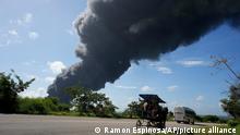 A carriage and bus drive past a plume of smoke caused by a blaze at the Matanzas Supertanker Base, in Matazanas, Cuba, Saturday, Aug. 6, 2022. Cuban authorities say lightning struck a crude oil storage tank at the base, causing a fire that led to four explosions which injured more than 50 people. (AP Photo/Ramon Espinosa)