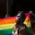 Pessoa negra em frente a uma bandeira do arco-íris. Ela está séria. 