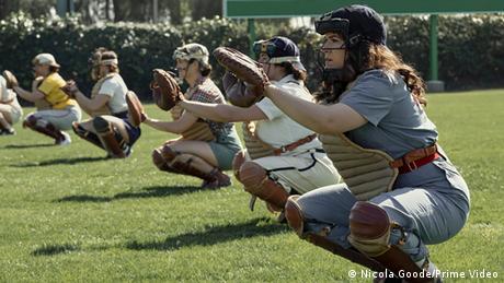 Six women, dressed in baseball jerseys and helmets with baseball gloves, squat on the playing field