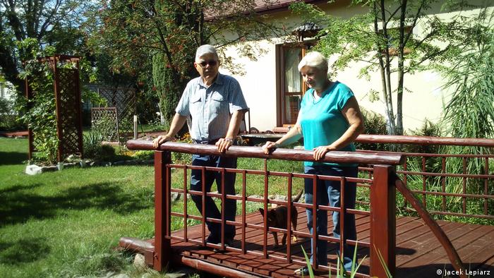 Das Rentner-Paar Barbara und Czeslaw Geszczak steht mit einem Hund auf der Holzterrasse vor seinem Haus.