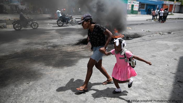 A woman guides a child past a demonstration amid the smoke of a street.