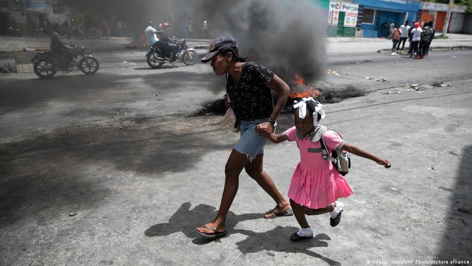 A woman guides a child past a demonstration amid smoke in a street.