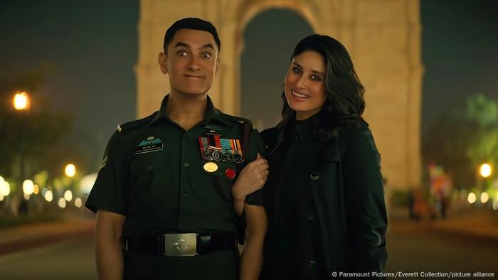 Ein Mann in einer Militäruniform und eine Frau in schwarzer Kleidung stehen vor einem Triumphbogen, sie lächelt in die Kamera.