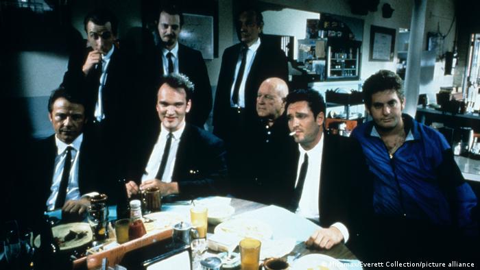 In a scene from the movie Reservoir Dogs, seven men in suits sit and stand around a table.