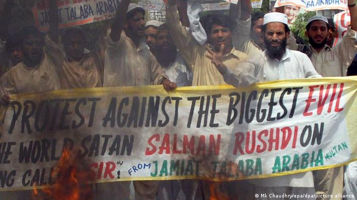 Wütende Männer halten ein Banner gegen den Satan Salman Rushdie, vor ihnen schlagen Flammen und Qualm hoch.