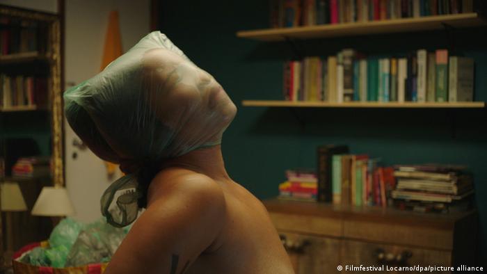 Filmszene: eine nackte Frau mit einer Tüte über dem Kopf, zu sehen sind Kopf und Schultern, im Hintergrund ein Bücherregal. 