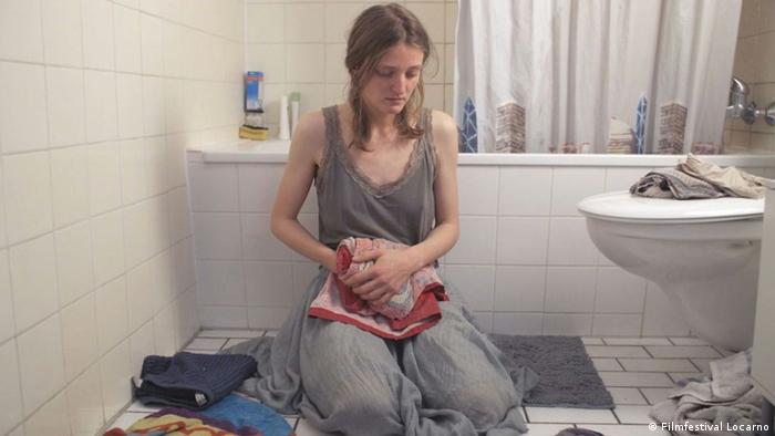 Filmszene: Eine Frau sitzt in einem grauen Kleid auf dem Fußboden in einem Badezimmer, sie blickt auf den Boden, in der Hand ein rotes Handtuch. 