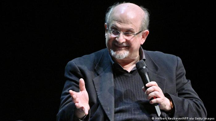 Salman Rushdie hält ein Mikrofon in der Hand und gestikuliert lächelnd.
