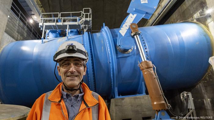 Das Bild zeigt den Kraftwerkdirektor vor einer großen, blauen Leitung im Kraftwerk. Er trägt einen Schutzhelm und einen organgefarbenen Pullover. 