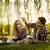 Pamje ilustruese - një vajzë dhe një djalë bëjnë piknik në park, të qeshur me një gotë verë para dhe kanistër me ushqime