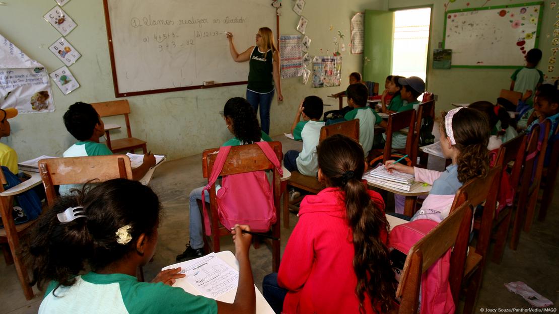 Crianças vistas de costas, diante de professora em sala de aula