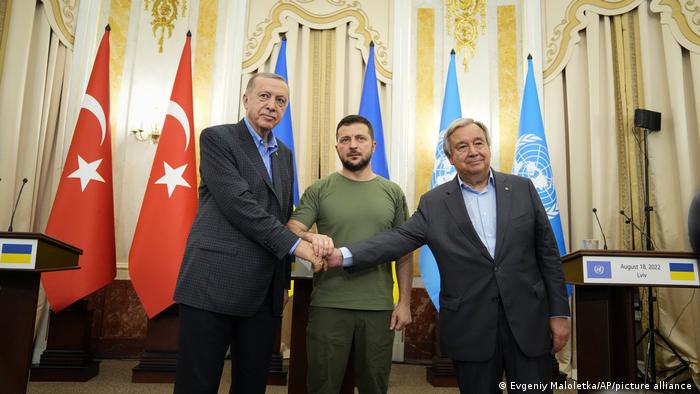 Handschlag von Erdogan, Selenskyj und Guterres vor den Fahnen der Türkei, der Ukraine und der UN