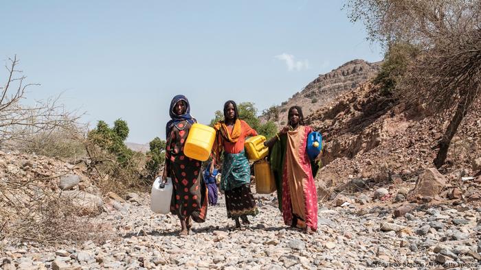 Mujeres caminan por un paisaje seco y pedregoso con garrafas de agua bajo el brazo.