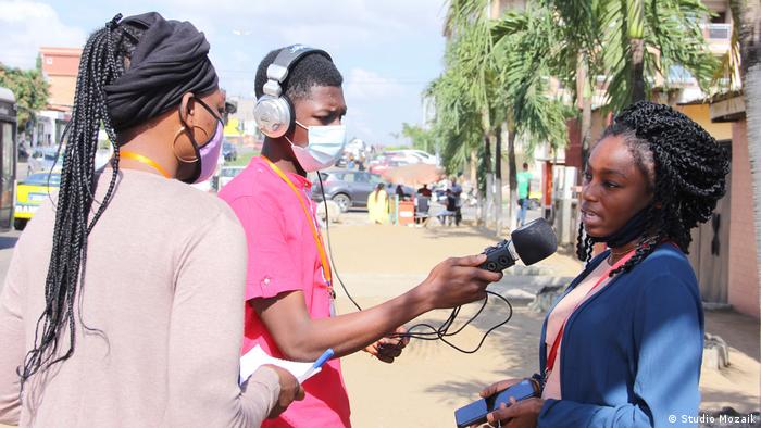 Two reporters interview a woman in the street using an audio recording device with a build-in microphone. The two reporters wear masks as protection against the coronavirus.