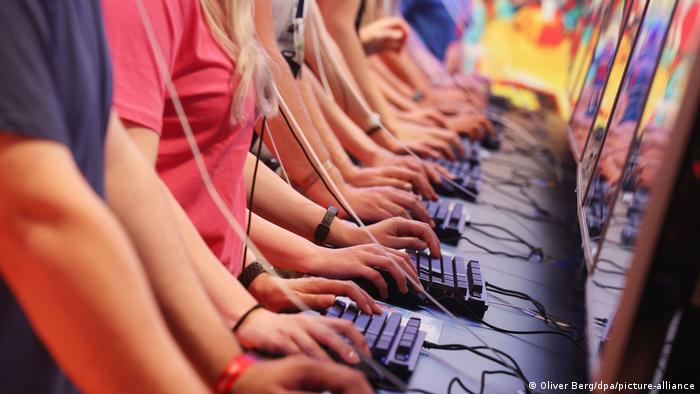 Gros plan sur les torses et les mains de plusieurs personnes, debout côte à côte, utilisant une rangée d'ordinateurs et de claviers tout en testant un jeu à la Gamescom.  24 août 2022. 