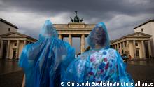 26.08.2022, Berlin: Ein Mann und eine Frau stehen mit Plastikregenschutz vor dem Brandenburger Tor. Foto: Christoph Soeder/dpa +++ dpa-Bildfunk +++