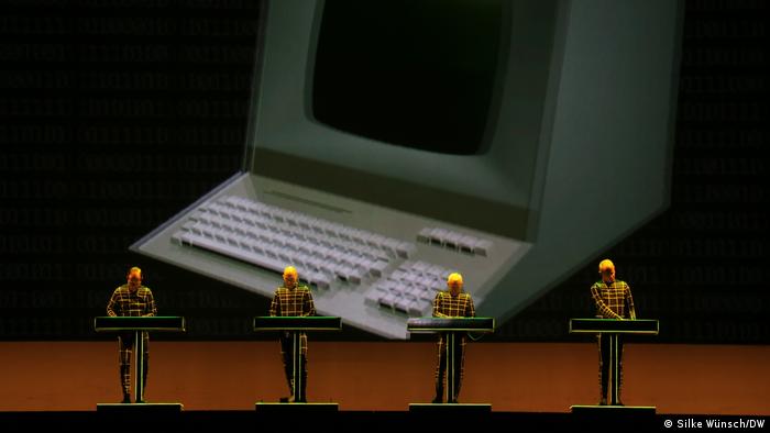 Kraftwerk live in Bonn: four men standing side by side at consoles, behind them the image of an early computer on a video screen.