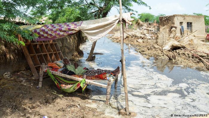 An old man lies on a bed in front of the remains of his house, surrounded by floodwaters