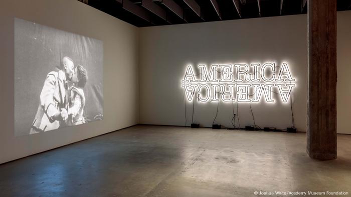 People kiss in a projection on one wall while on another the word America is illuminated