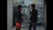 Filmstill aus 'Black Chariot': ein Mann und eine Frau stehen vor einer Hauswand mit Postern und unterhalten sich Filmstill aus 'Black Chariot': ein Mann und eine Frau stehen vor einer Hauswand mit Postern und unterhalten sich