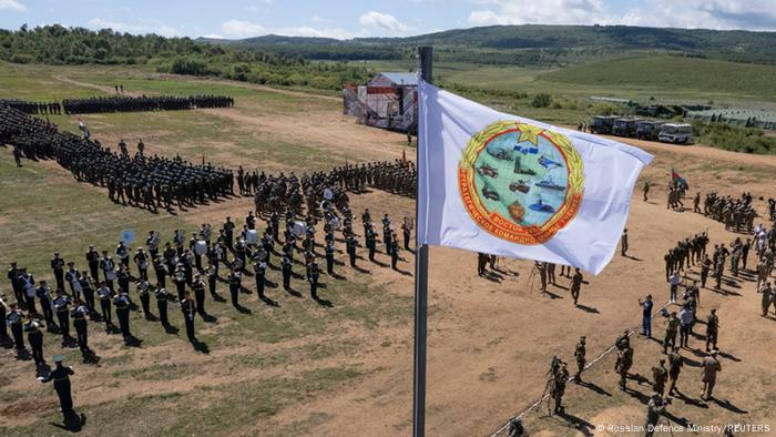 Über einem Platz, auf dem Soldaten in Formationen stehen, weht eine weiße Fahne mit der Abbildung von militärischen Fahrzeugen im Wind. Feierliche Eröffnung einer gemeinsamen Militärübung von Russland, Indien und China im August 2022 in der russischen Region Primorje