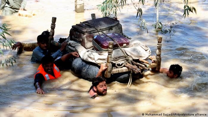 Menschen in Pakistan waten mit gerettete Habseligkeiten auf einem Floß durch ein überschwemmtes Gebiet