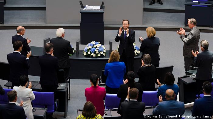 Blick in den Plenarsaal des deutschen Bundestags. Ein Mann mit zum Applaus erhobenen Händen steht vor mehreren Personen mit dem Rücken zum Betrachter: Sie haben sich von ihren Stühlen erhoben und klatschen ihm Beifall