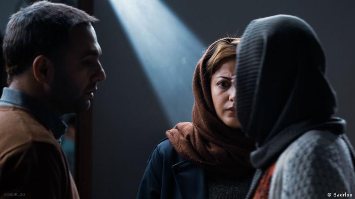 Film scene from Bi Roya, a man stands in the shadow on the left and looks seriously at two women wearing headscarves