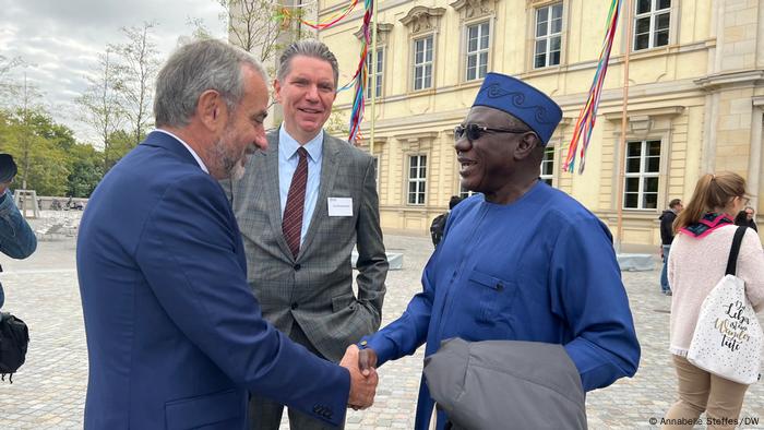 Hermann Parzinger und Abba Isa Tijani schütteln einander die Hand. Sie tragen blau und stehen vor dem Stadtschloss in Berlin.