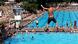 A boy is seen mid-air as he jumps off a diving block. Below him the pool and pool grounds are crowded with people. A boy is seen mid-air as he jumps off a diving block. Below him the pool and pool grounds are crowded with people.