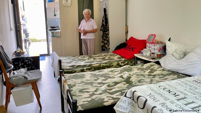 Eine ältere Frau steht in einem Wohncontainer, in dem zwei Betten und ein Schrank aufgestellt sind. Im Vordergrund ist ein Tisch zu sehen, links an der Wand ein Stuhl. Im Hintergrund ist der Ausgang und die Kochnische