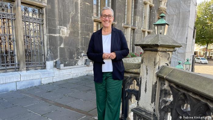 Oberbürgermeisterin Sibylle Keupen steht vor dem Rathaus in Aachen. Sie trägt eine grüne Hose, ein weißes Shirt und eine schwarze Jacke. Ihre grauen Haare sind kurz geschnitten und sie trägt eine Brille mit dunklem Rahmen. 