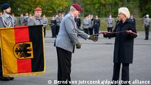 Christine Lambrecht übergibt dem Generalinspekteur der Bundeswehr, Eberhard Zorn, ein Fahnenband. Links im Bild die schwarz-rot-goldene deutsche Flagge.