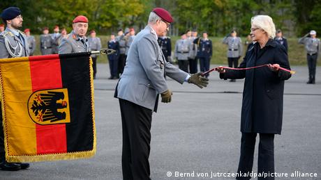 Christine Lambrecht übergibt dem Generalinspekteur der Bundeswehr, Eberhard Zorn, ein Fahnenband. Links im Bild die schwarz-rot-goldene deutsche Flagge.